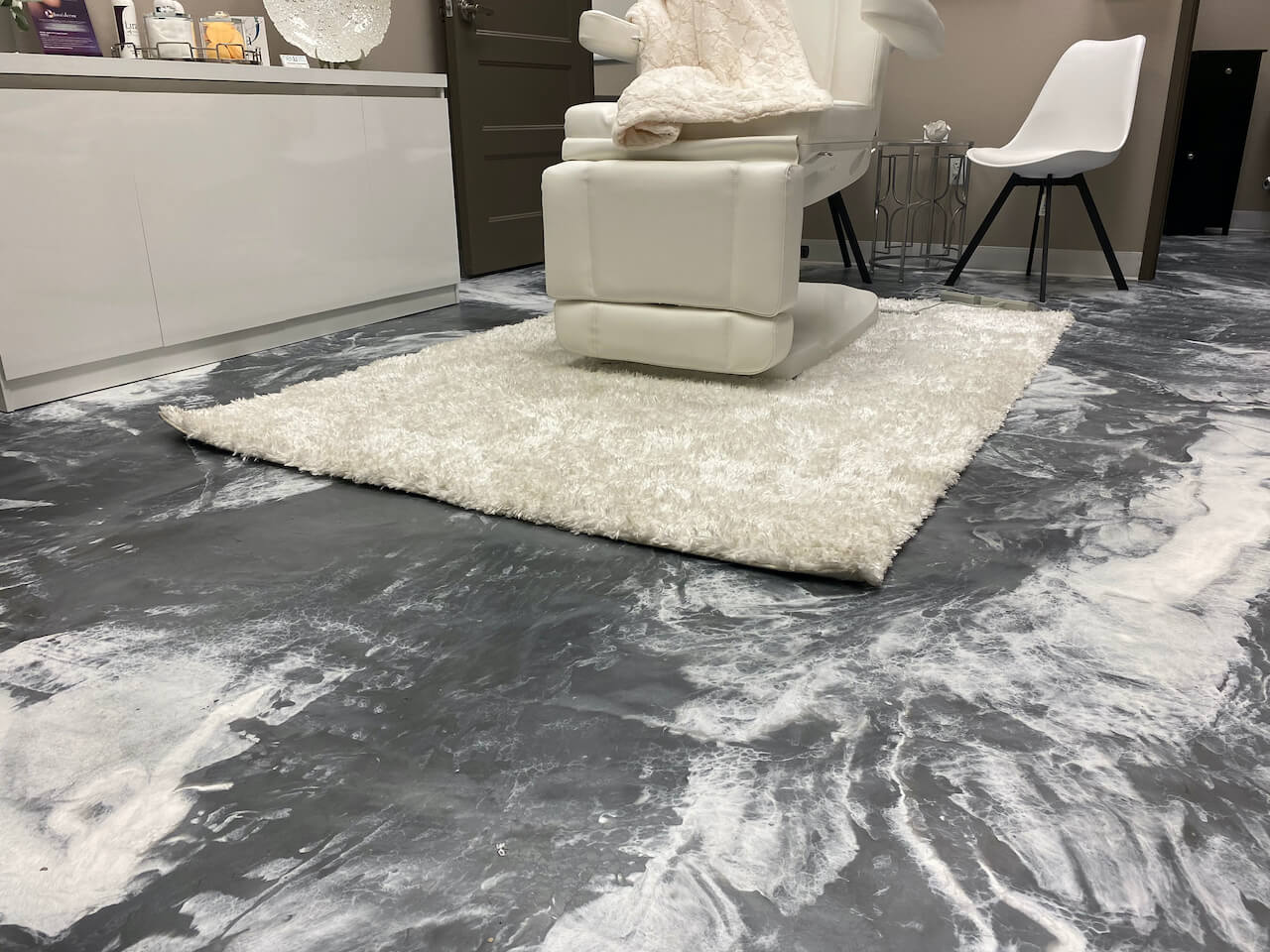 Dark gray and white metallic epoxy floor with a white rug and chair on top.  The floor mimics real stone.